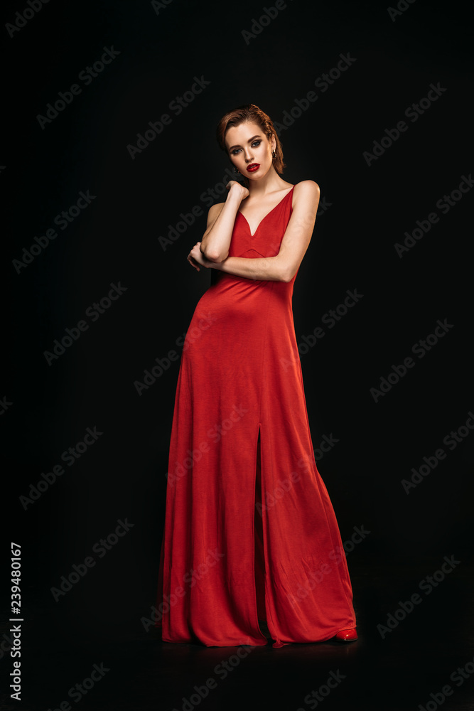 low angle view of attractive girl in red dress and black jacket looking at camera isolated on black