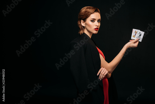 side view of attractive girl in red dress and black jacket holding poker cards and looking at camera isolated on black