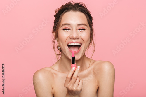 Pretty happy young woman posing isolated over pink wall background holding lipstick.