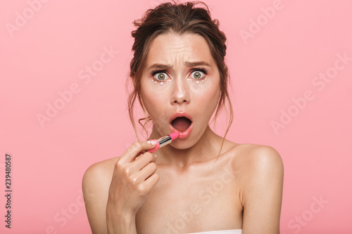 Shocked young woman posing isolated over pink wall background holding lipstick.