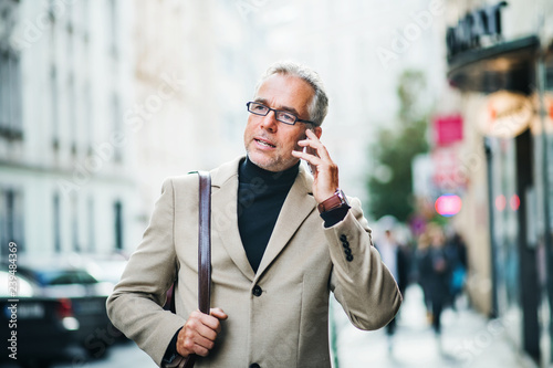 Sticker Mature businessman standing on a street in city, using smartphone