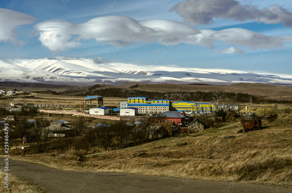 Winter afternoon with huge clouds on the blue Sunny sky over a mountain village with 
 enterprise in the lowland of the snow-covered mountains of Aragats