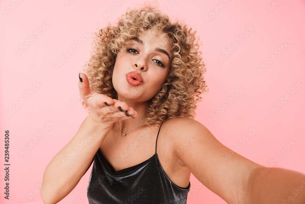 Portrait of stylish curly woman 20s smiling and taking selfie photo, isolated over pink background