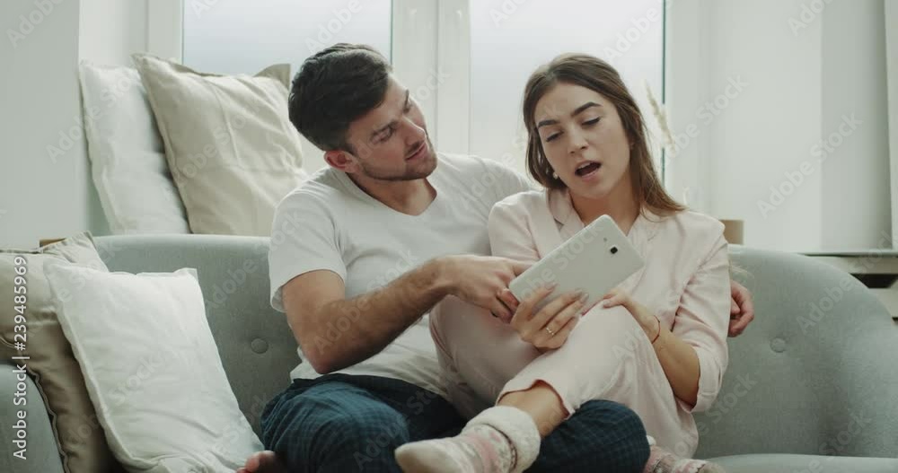 Using a tablet couple order something from phone they are in pajamas sitting on sofa and have a happy mood. Using a tablet couple order something from phone they are in pajamas sitting on sofa and have a happy mood.