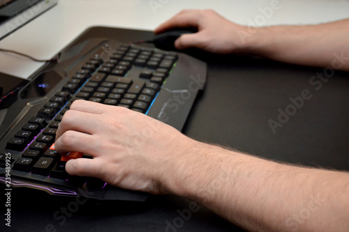 Close-up of male hands on a black gaming keyboard with neon light on a black table. Side view.