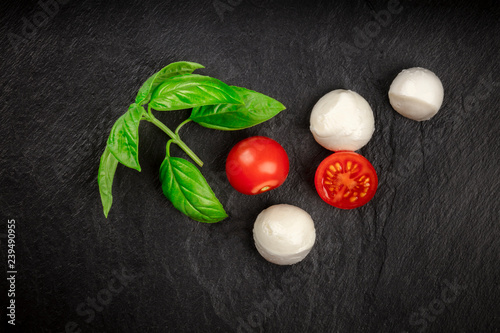 An overhead photo of Mozzarella cheese balls with fresh basil leaves and cherry tomatoes, the ingredients of the Italian Caprese salad, on a black background with a place for text