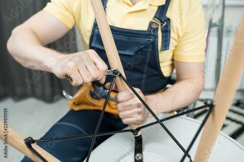 Close-up of unrecognizable handyman in denim overall using wrench while repairing chair in apartment