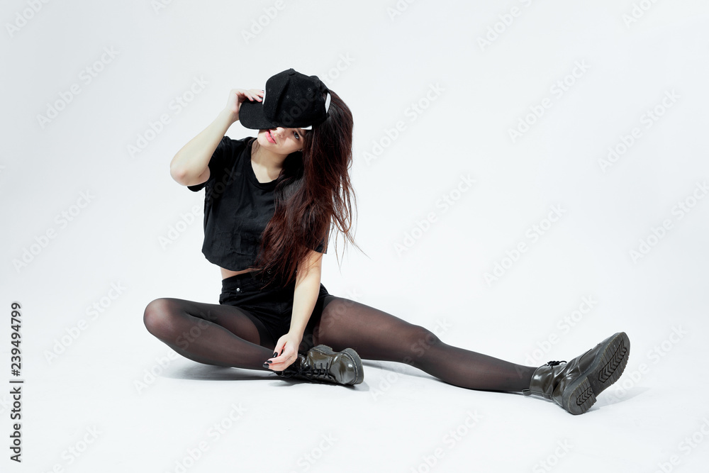 Funny young stylish girl dressed in a black top, shorts, tights and cap  sits on the floor  on the white background in the studio