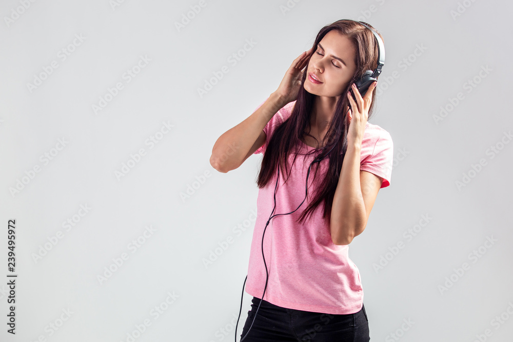 Charming girl dressed in pink t-shirt and jeans is listening music in the headphones and with the phone in her hand on the white background in the studio Charming girl dressed in pink t-shirt and jeans is listening music in the headphones and with the phone in her hand on the white background in the studio