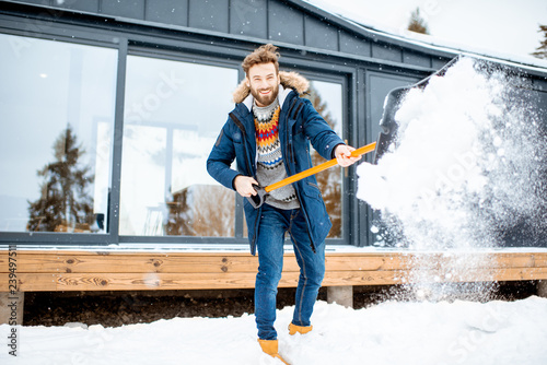 Handsome man in winter clothes cleaning snow with a shovel near the modern house in the mountains