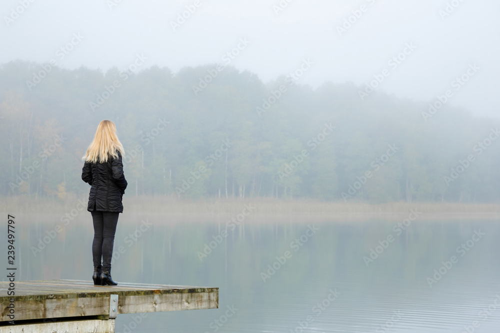 Young adult blonde woman standing alone on edge of footbridge and staring at lake. Mist over water. Foggy air. Early chilly morning. Peaceful atmosphere in nature. Back view.