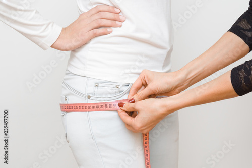 Woman's hands measuring young, slim girl's hips with pink measuring tape. Part of body on gray background. Right size. Closeup. Side view. 