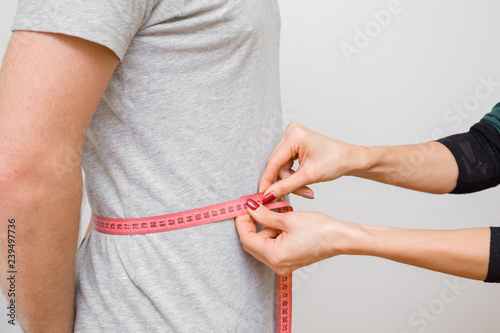 Woman's hands measuring young, slim man's waist with pink measuring tape. Part of body on gray background. Right size. Closeup. Side view. photo