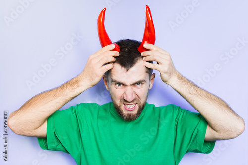 Young man holding red capi peppers above his head which looks like horns, angry and evil face. Light purple background. photo