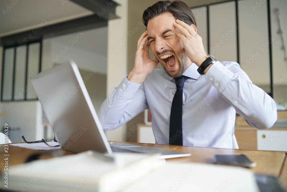 Angry businessman losing patience and screaming at laptop in modern office Angry businessman losing patience and screaming at laptop in modern office