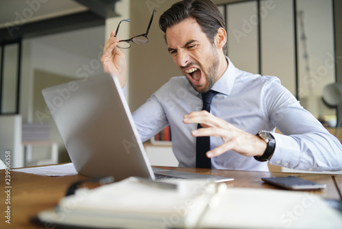 Angry businessman losing patience and screaming at laptop in modern office