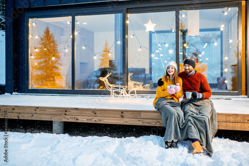 Young couple warming up with plaid and hot drinks sitting on the terrace of the modern house in the mountains durnig the winter holidays