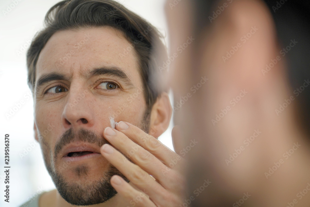 Obraz premium Handsome man applying face cream looking in mirror