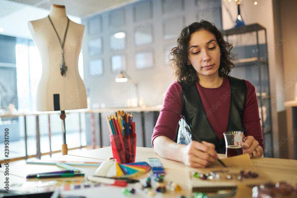 Fototapeta premium Portrait of creative young woman drawing sketches for jewelry while working at table in sunlit shop, copy space