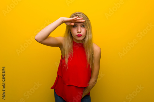 Young girl with red dress over yellow wall looking far away with hand to look something
