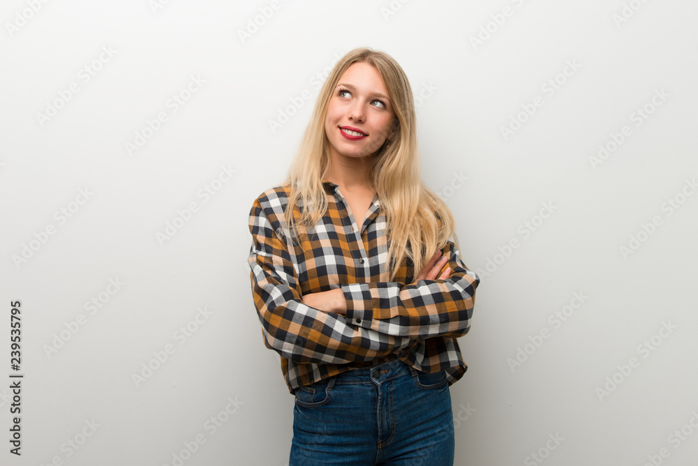 Blonde young girl over white wall looking up while smiling Blonde young girl over white wall looking up while smiling