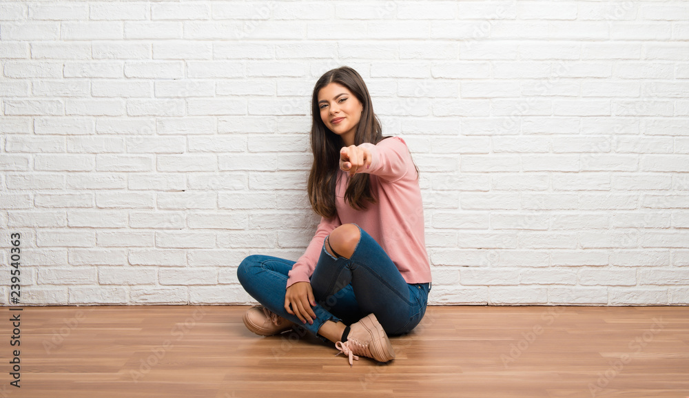Teenager girl sitting on the floor in a room points finger at you with a confident expression Teenager girl sitting on the floor in a room points finger at you with a confident expression