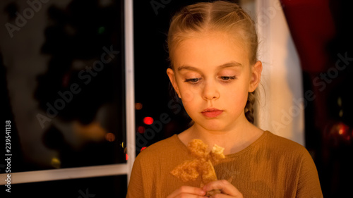 Sad little girl waiting for parents near orphan home window  eating Xmas cookie