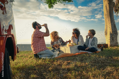 Friends eat on meadow happy young blond and curly woman with plaid shirt plays guitar brunette friend with headband smiles singing man with beard drinks bottle of beer and blond child looks amused