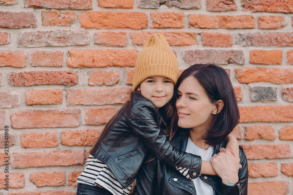 Affectionate mother with dark hair recieves warm hug from daughter, wear leather jackets, pose together against brick wall. Fashionable little girl in hat embraces her mum. Family and children concept