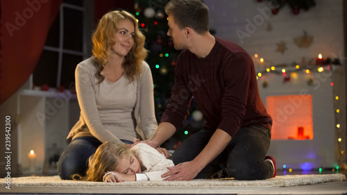 Loving child fallen asleep under Christmas tree, happy parents sitting beside