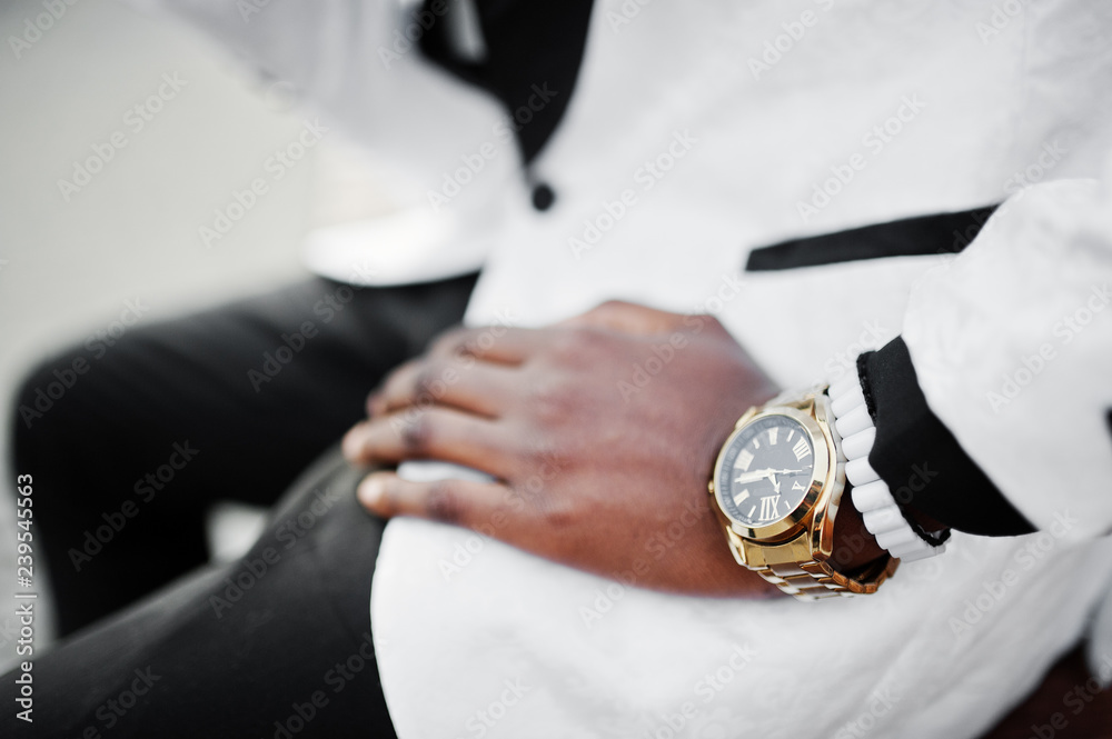 Obraz premium Chic handsome african american man in white suit sitting on bench. Close up photo of golden watches on hand.