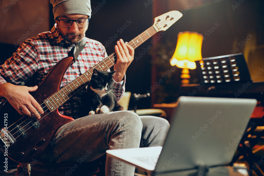 Obraz premium Young Caucasian man with hat on head playing bass guitar while sitting in home studio.