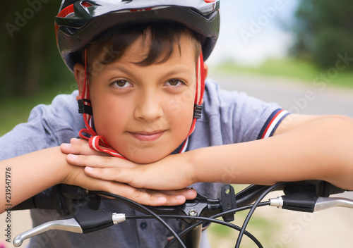 Foto Close-up of a teenager boy in protective helmet sitting on his bicycle and keeps his head on his hands resting on the handlebars of the bike in park on summer day