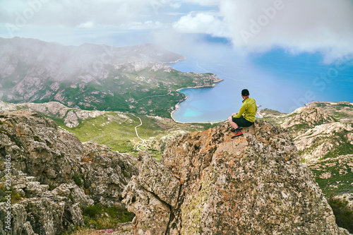 Trourist boy sitting on the rock mountain high above the sea and looking down Corsica Corse France