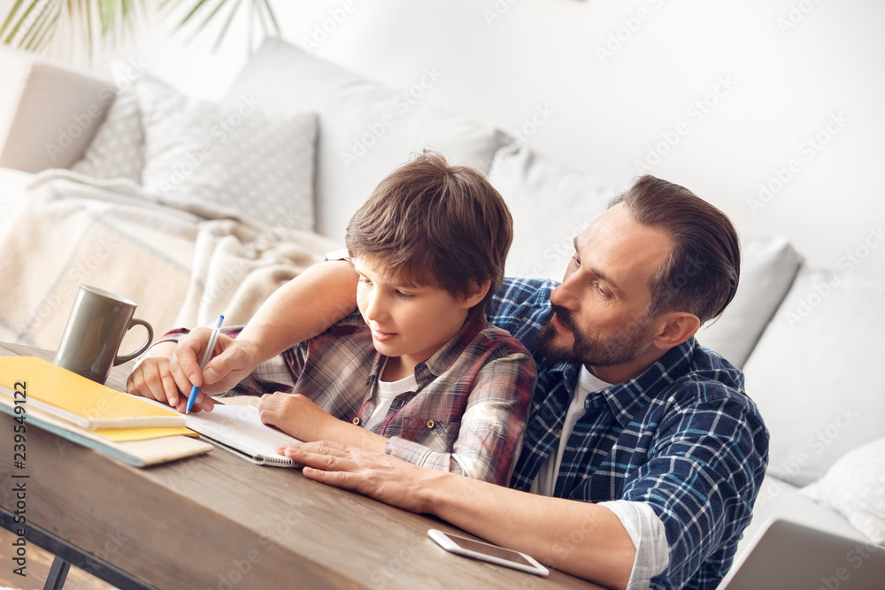 Father and son at home sitting at table dad holding boy's hand writing assignment together in notebook Father and son at home sitting at table dad holding boy's hand writing assignment together in notebook