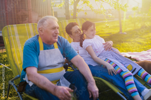 Older couple sitting with their granddaughter in backyard and making barbeque. Family time.