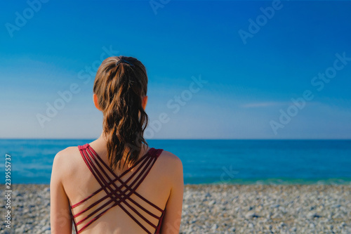 Woman wearing yoga top with cross strips back. Girl meditating on a pebble beach by the sea at sunny day with clear blue sky.
