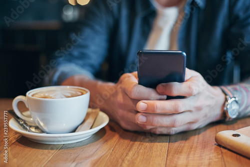 Man hands in denim shirt slide with finger on screen his smart phone, near cup with coffee on wooden table