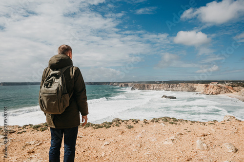 Rear view of a man tourist or a guy with a backpack admires the beautiful landscape and the ocean in solitude and thinks or dreams.