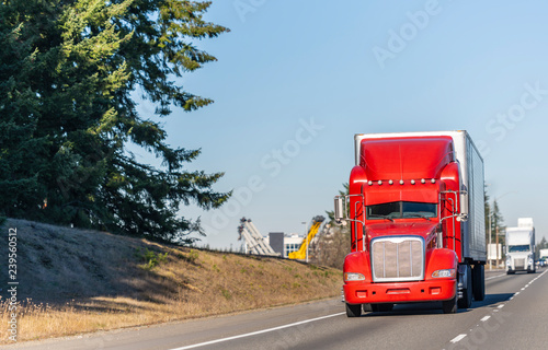 Big rig bright red classic American idol semi truck with reefer semi trailer driving on the straight highway in sunny day
