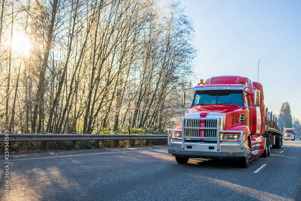 Obraz Big rig modern red semi truck with flat bed semi trailer transporting commercial cargo on the road with sun shining through the trees