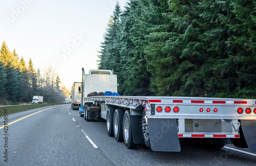 Big rig long haul semi truck with empty flat bed semi trailer running on the winter road in convoy with another semi trucks