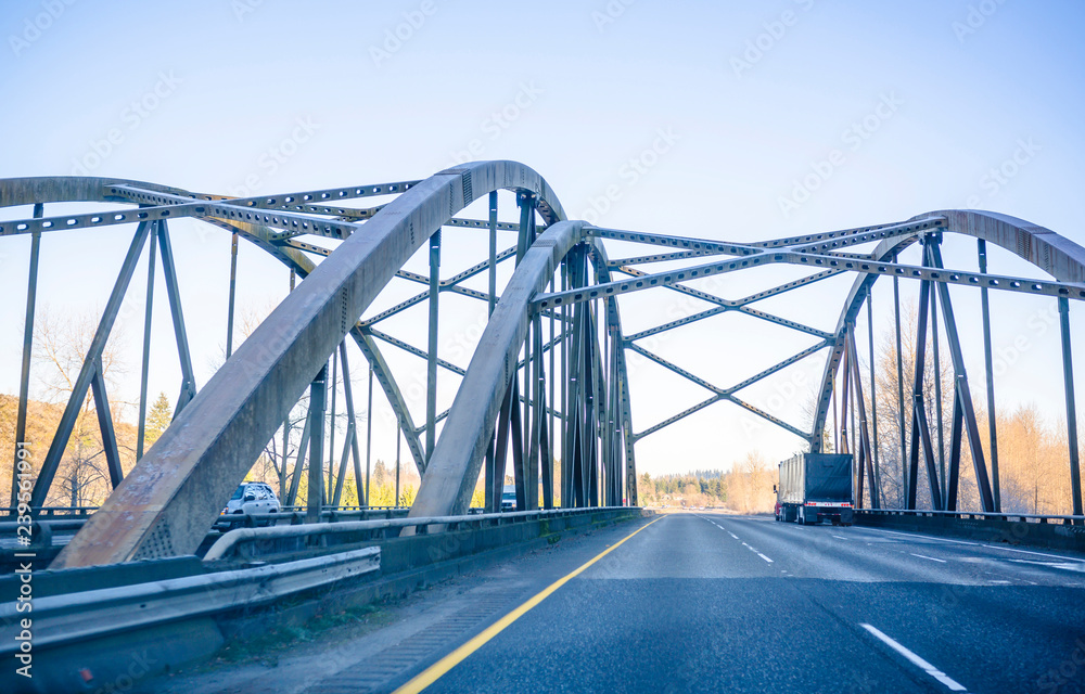Big rig semi truck transporting covered semi trailer driving on the winter frosty arched truss bridge