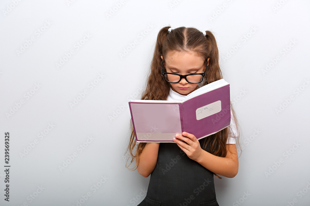 Smart pupil kid girl in eyeglasses looking in the textbook and dooing the exercices on blue color background Smart pupil kid girl in eyeglasses looking in the textbook and dooing the exercices on blue color background