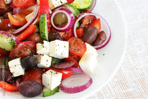Greek Salad of tomatoes, cucumbers, sweet peppers, feta cheese, dark olives, purple onions on white plate. Closeup