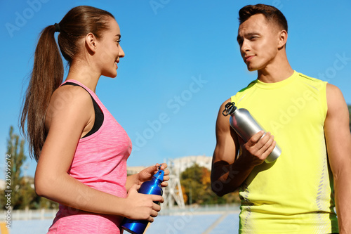 Young sporty couple holding bottles of water at stadium on sunny day