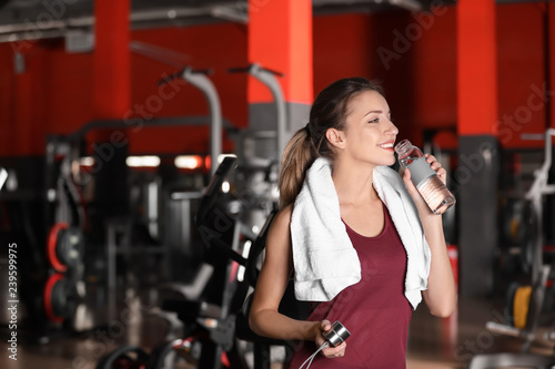 Beautiful young woman with towel and bottle of water in gym. Space for text