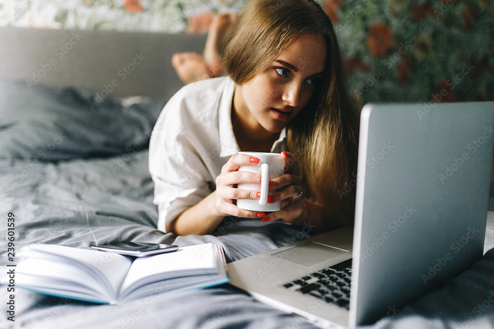 Attractive smiling european woman using laptop and drinking coffee while lying on her bed. Young woman drinking coffee at home, start, concept in the morning, early in the morning. Stylish Toned Attractive smiling european woman using laptop and drinking coffee while lying on her bed. Young woman drinking coffee at home, start, concept in the morning, early in the morning. Stylish Toned