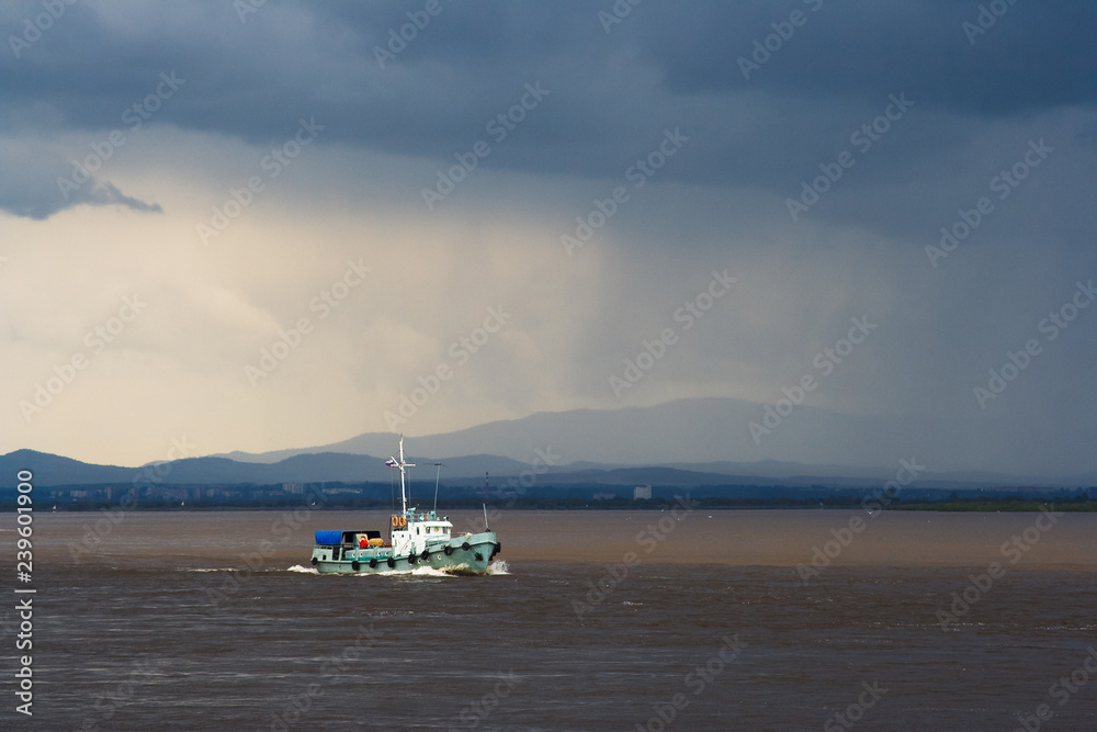Landscape with a large river, which floats boat. In the sky dark clouds. Gloomy weather, it is raining. Amur River, Khabarovsk Region, Far East of Russia.