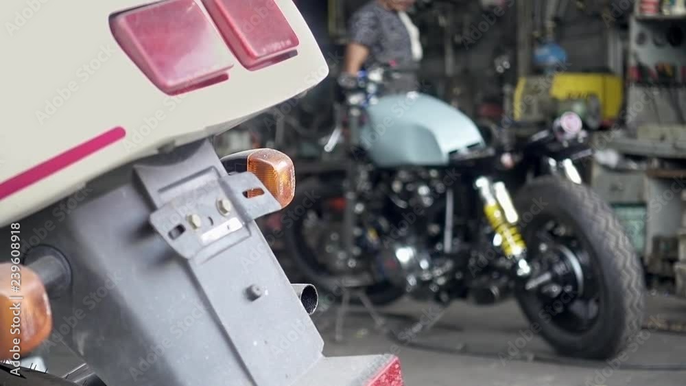 Shot of a mechanician plan to work with his motorcycle as blurry background, taillight of white motorcycle in foreground. Handheld slow-motion shot, 29.97fps. Shot of a mechanician plan to work with his motorcycle as blurry background, taillight of white motorcycle in foreground. Handheld slow-motion shot, 29.97fps.